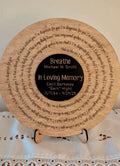 A circular wooden memorial plaque displayed on a small stand, featuring engraved text around the edge and a black center with personalized remembrance text.
