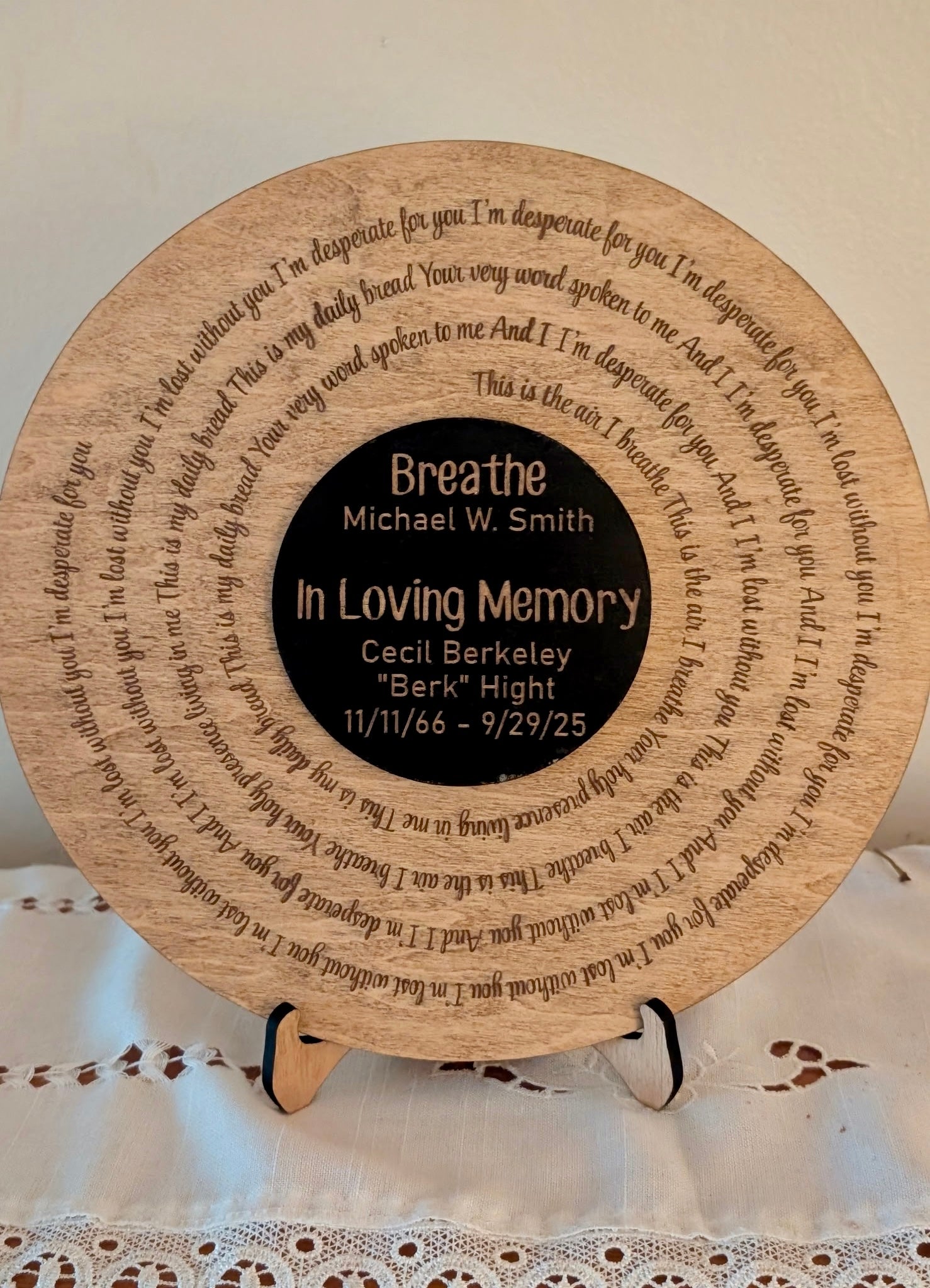 A circular wooden memorial plaque displayed on a small stand, featuring engraved text around the edge and a black center with personalized remembrance text.