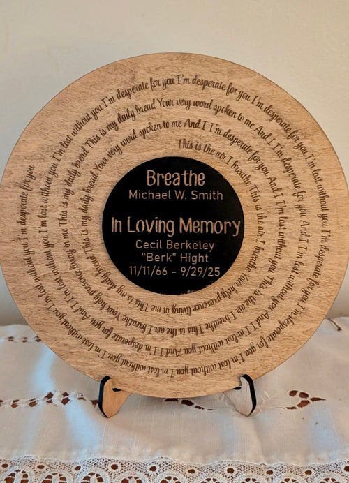 A circular wooden memorial plaque displayed on a small stand, featuring engraved text around the edge and a black center with personalized remembrance text.